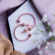 A pair of rose gold semi-hoop earrings with a textured surface, displayed on a white surface with a blurred background featuring a purple satin drape and flowers.